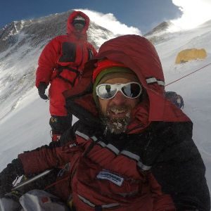 Photo of Jim Davidson in the foreground and his Mount Everest Guide in the background sitting on Mount Everest with yellow tents in the background